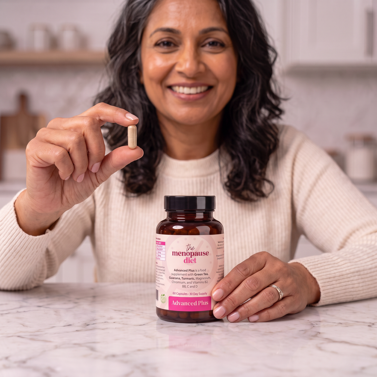 A woman in a cream knit sweater holds up a single supplement capsule next to a brown bottle labeled 'The Menopause Diet' on a white marble countertop in a bright kitchen.